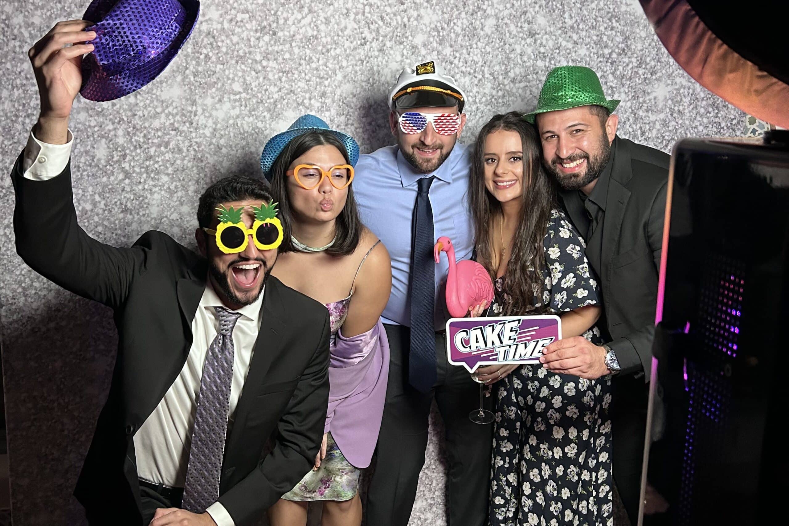 Group of five friends posing in a photo booth, wearing colorful hats and oversized sunglasses, holding a "Cake Time" sign, celebrating at an event.