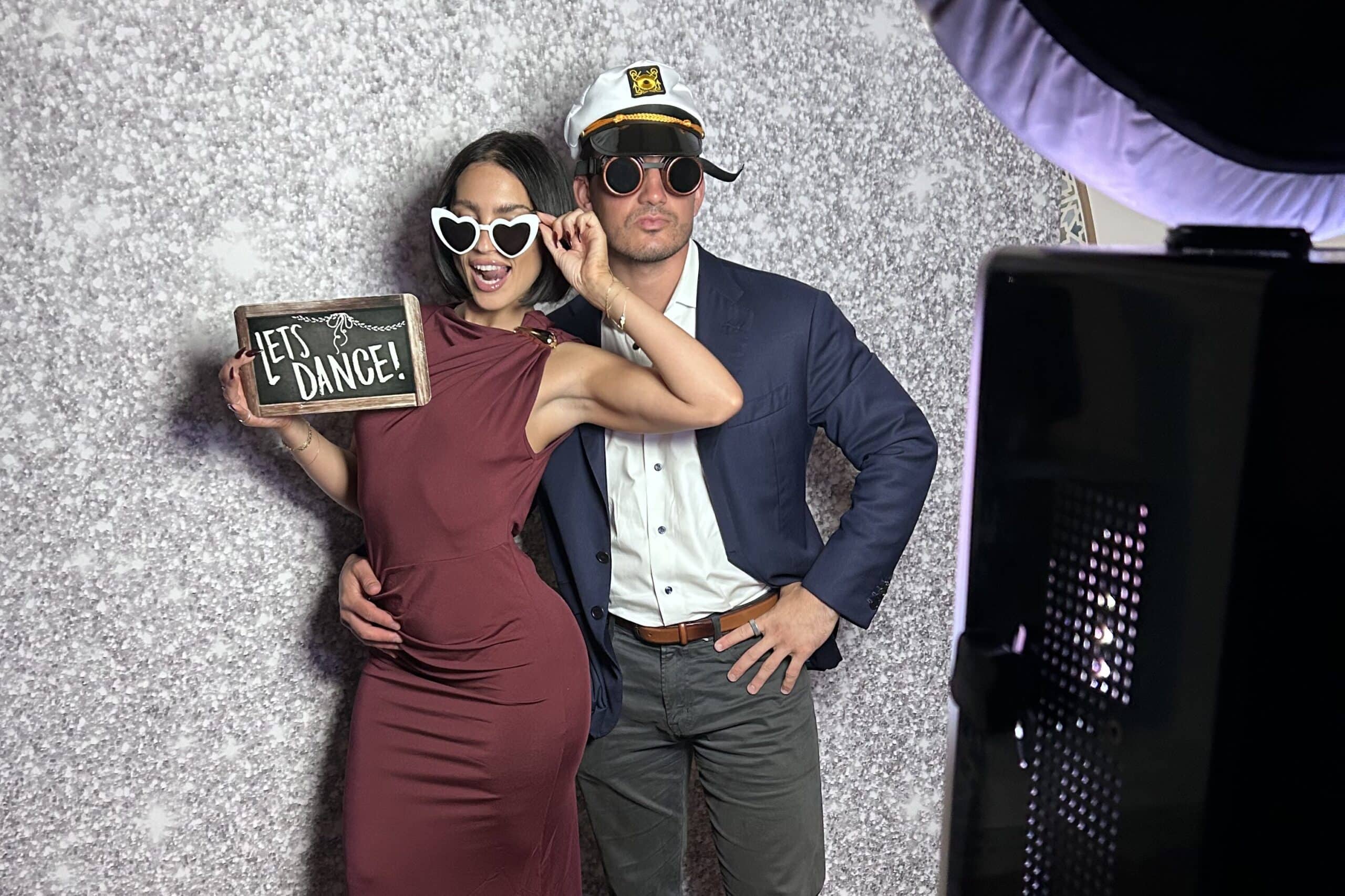 Couple posing in a photo booth, wearing fun sunglasses and a captain's hat, holding a sign that says "Let's Dance!" against a sparkly backdrop, illustrating a lively photo booth experience for social media sharing.