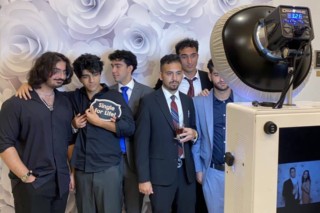 Group of young men posing in front of a floral backdrop, holding a humorous sign reading "Single for Life," with a photo booth capturing the moment, illustrating interactive engagement at a social event.